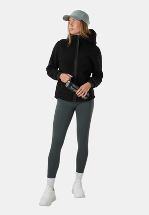 Kvinde iført sort fleecejakke, grå leggings, hvide sneakers og lysegrøn kasket, holder en sort vandflaske og kigger til siden.