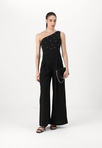 Black one-shoulder jumpsuit with pearl embellishments, wide-legged design, paired with a pleated black clutch and strappy sandals.
