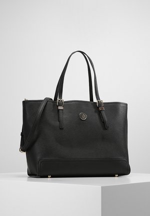 Sac tote structuré noir avec double bandoulière, sangle bandoulière réglable et petit logo métallique rond à l'avant, posé sur une surface blanche.