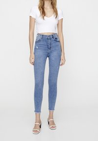 Jean bleu taille haute avec une finition déchirée et un ourlet brute. Associé à un t-shirt court blanc ajusté et des sandales à talons blanches.