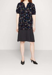 Svart blommig blus med korta puffärmar över en mörk kjol. Tyget har färgglada, små blommönster. Svarta högklackade mules.