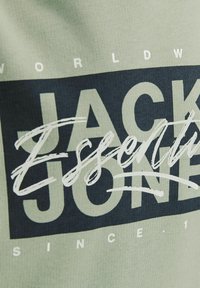 T-shirt en coton vert avec une impression navy audacieuse, avec "JACK & JONES" et "Essentials" en script blanc et une texture côtelée.