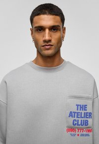 Grijze sweatshirt met een voorzak en blauwe en rode tekst. Zachte textuur en een relaxte pasvorm met een ronde halslijn en zichtbare stiksels.