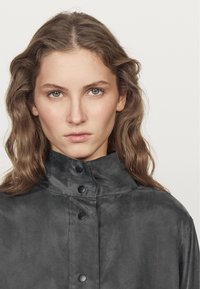 Gray, high-collar jacket features a smooth texture and black snap buttons along the front. Minimalist design with a structured silhouette.
