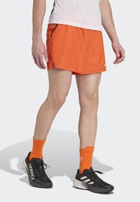 Pantalones cortos deportivos naranjas fabricados en tejido ligero con aberturas laterales y con detalles en negro, combinados con zapatos negros y calcetines naranjas.