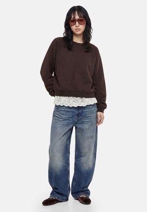 Persona che indossa jeans blu oversize, felpa marrone, top bianco di pizzo che sbuca, ballerine di velluto marrone e grandi occhiali da sole marroni, in piedi su sfondo bianco.