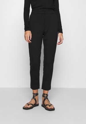Personne portant un haut noir à manches longues, un pantalon noir longueur cheville et des sandales noires avec brides et clous, debout devant un fond uni.