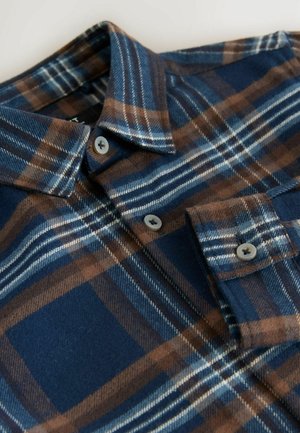 Chemise en flanelle bleu marine avec un motif à carreaux marron et blanc. Présente un col boutonné et des boutons gris. Texture douce visible.