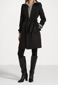 Svart trenchcoat med dubbelknappning, bälte i midjan och axelkläppar. Buren över en randig skjorta och i kombination med svarta knähöga stövlar.