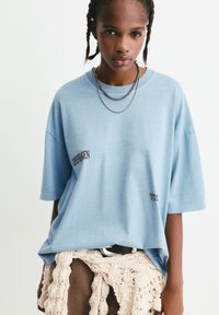 Junge Person mit geflochtenem Haar, die ein hellblaues, übergroßes T-Shirt mit Text, geschichtete Halsketten, braune Hosen und ein cremefarbenes gestricktes Kleidungsstück, das um die Taille gebunden ist, trägt.