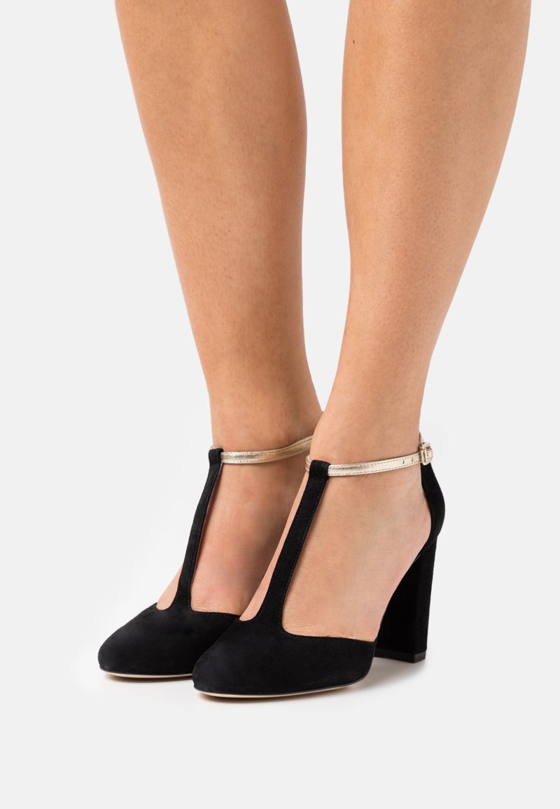 Chaussures à talons hauts en daim noir avec un design à lanière en T, dotées d'une bride de cheville métallique dorée et d'un talon carré. Texture lisse avec des découpes.