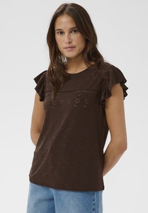 Mladá žena s tmavohnedými vlasmi, oblečená v hnedej blúzke s dierkovaným vzorom a volánikovými rukávmi a svetlomodrých džínsoch, na jednoduchom pozadí.