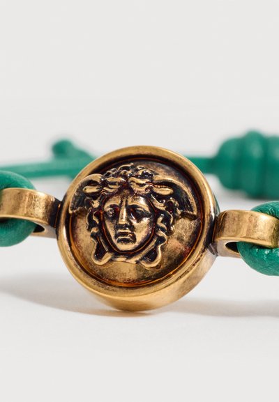 Médaillon en or avec un motif de tête de Méduse attaché à un bracelet en cuir vert sur fond blanc.