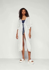 Kimono trasparente bianco con un motivo a zig-zag, dotato di maniche ampie e chiusura a nodo in vita, abbinato a un costume da bagno blu navy.