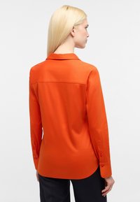 Camicia a maniche lunghe in tessuto arancione, con colletto, cucitura della spalla sul retro e polsini abbottonati, caratterizzata da una texture liscia e una vestibilità aderente.