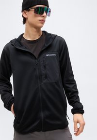 Νεαρός άνδρας που φορεί μαύρο σκουφί, μεγάλα ημιδιάφανα γυαλιά ηλίου και μαύρο μπουφάν Columbia με φερμουάρ, με χέρια στις τσέπες, μπροστά σε απλό φόντο.