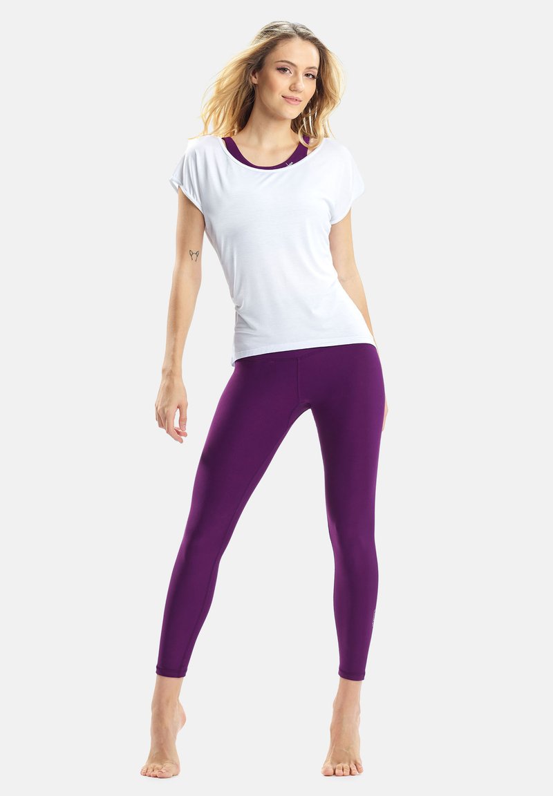 Fehér rövid ujjú felső, amelyhez testhezálló lila leggings tartozik. A leggings sima textúrájú, és mindkét darab modern, sportos szabású.