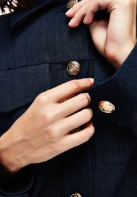 Tummansininen farkkutakki, jossa on kullanväriset koristeelliset napit, kaksi rintataskua ja napitettava kaulus. Keskittyminen takin napittamiseen käsin.