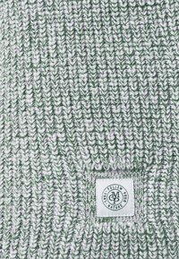 Bonnet en tricot vert et blanc, avec un motif texturé. Une petite étiquette blanche avec du texte noir est visible sur le côté.
