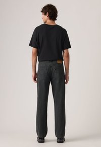 Μαύρο κοντομάνικο μπλουζάκι συνδυασμένο με σκουρόχρωμο γκρι, υφασματώδες τζιν με κάθετες ρίγες και πίσω τσέπες. Το μοντέλο στέκεται με την πλάτη προς τον θεατή.