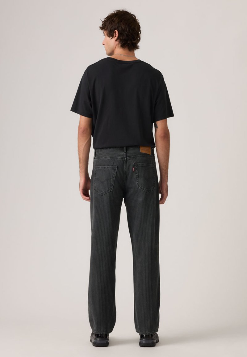 T-shirt noir à manches courtes associé à un jean gris foncé texturé avec des rayures verticales et des poches arrière. Le modèle est debout, dos tourné vers le spectateur.