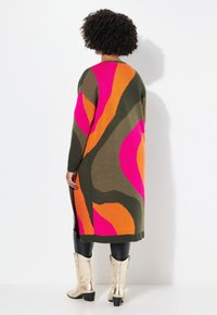 Long cardigan en tricot arborant des motifs abstraits audacieux dans des teintes de rose, orange et vert olive. Manches et ourlet côtelés, associés à des bottes dorées brillantes.