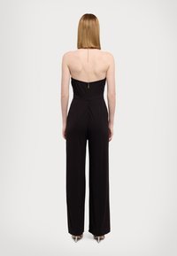 Combinaison noire au design sans bretelles, fermeture éclair dans le dos, avec un détail de chaîne en or le long du dos. Tissu doux et lisse, jambes larges.