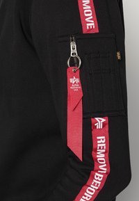 Felpa nera con tasca con zip e strap rosso con testo "RIMUOVERE PRIMA". Tessuto testurizzato con accenti a contrasto.