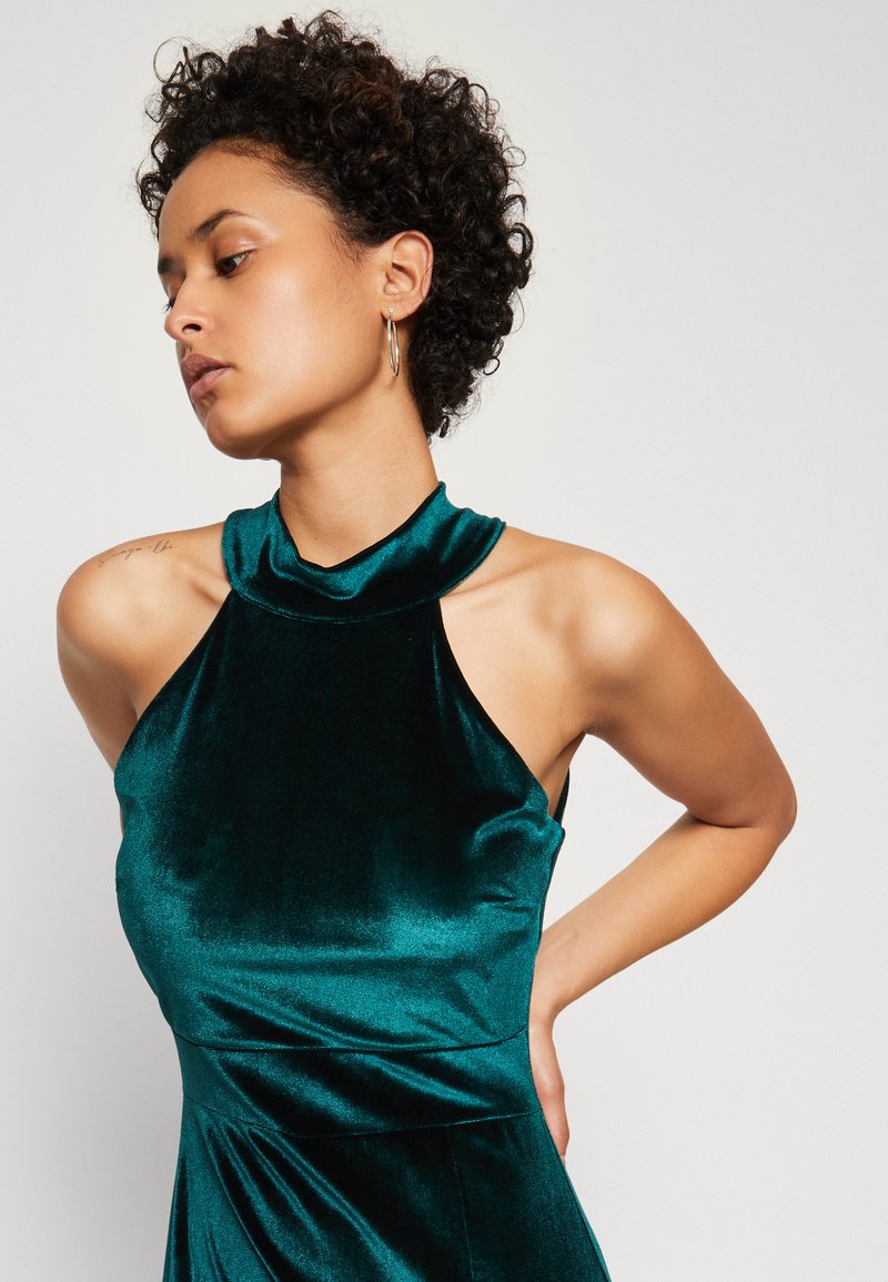 Abito in velluto color teal con colletto alto e senza maniche. Presenta una consistenza soffice, drappeggio elegante e un corpetto aderente con gonna svasata.