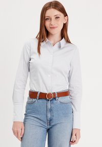 Chemise blanche à manches longues avec des rayures bleues. Associée à un jean bleu taille haute et à une ceinture marron avec une boucle ronde.
