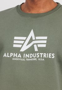 Ελιάς πράσινη φούτερ μπλούζα με στρογγυλή λαιμόκοψη, που διαθέτει μια μεγάλη λευκή γραφική λογότυπο και το όνομα της μάρκας "Alpha Industries" τυπωμένο στο κέντρο.