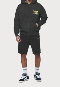 Musta vetoketjuhuppari, jossa on graafinen logo, yhdistettynä mustiin farkkushortseihin. Päällä valkoiset sukat ja tummansiniset lenkkitossut.