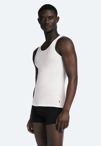 Hvid ribbet tanktop lavet af bomuld, tætsiddende design. Sort trusse nedenunder, båret af en person mod en lys grå baggrund.