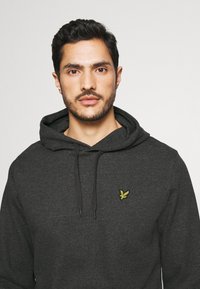 Haut à capuche gris charbon en tissu doux, avec une poche kangourou, des cordons de serrage réglables et un petit logo jaune sur la poitrine.