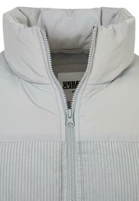 Lichtgrijs gewatteerd jack met hoge kraag, ritssluiting aan de voorkant en geribbelde details aan de onderkant.