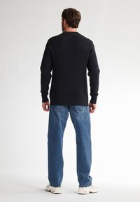 Zwarte, ribbelgebreide trui met lange mouwen; gecombineerd met lichtblauwe rechte jeans en witte sneakers. Zicht van achteren.