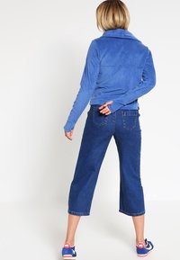 Giacca in pile blu con colletto alto e maniche lunghe, abbinata a pantaloni in denim larghi e cropped. Scarpe sportive con accenti blu e rosa.