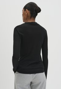 Frau mit dunklem Haar im Dutt, die ein schwarzes Langarmoberteil und hellgraue Hosen trägt, mit dem Rücken zu einem schlichten hellen Hintergrund.