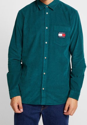 Turkusowa koszula sztruksowa z długim rękawem, zapinana na guziki, z kieszenią na piersi z logo Tommy Jeans, noszona z ciemnoniebieskimi jeansami.