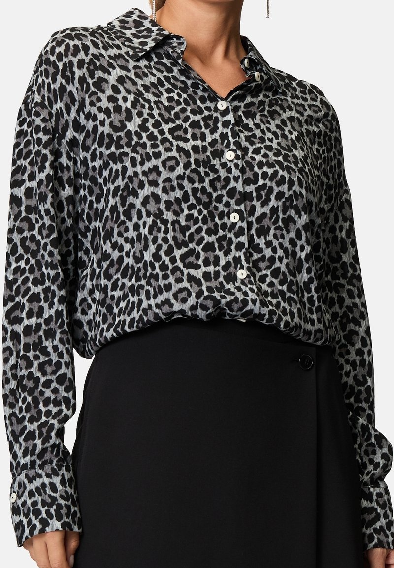Schwarz-graue Bluse im Leopardenmuster mit Kragen, Knopfleiste und langen Ärmeln, kombiniert mit einem schwarzen Rock mit seitlichem Knopf.