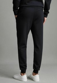 Pantaloni de trening negri dintr-un material moale, cu o croială ajustată, având buzunare laterale și un buzunar mic la spate, asortați cu adidași albi.