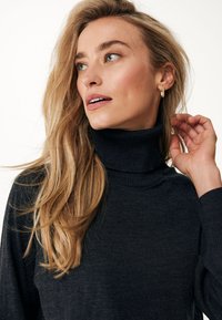 Maglione grigio scuro a collo alto con scollo a coste e maniche lunghe, caratterizzato da una morbida trama. Capelli acconciati in modo casuale, con piccoli orecchini a cerchio.