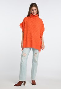 Poncho de punto naranja con cuello alto y bordes con flecos, combinado con jeans rotos de color azul claro y botas de tobillo marrones con tacón.