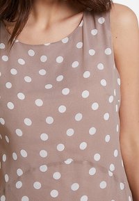 Hellbraunes, ärmelloses Top mit glatter Textur, das große, gleichmäßig verteilte weiße Polka-Dots auf dem Stoff zeigt. Rundhalsausschnitt.