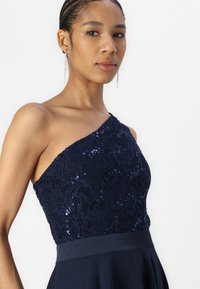 Robe bleu marine avec un design à une épaule, présentant un haut en dentelle à paillettes et un bas en satin lisse, rehaussée d'une ceinture en satin.