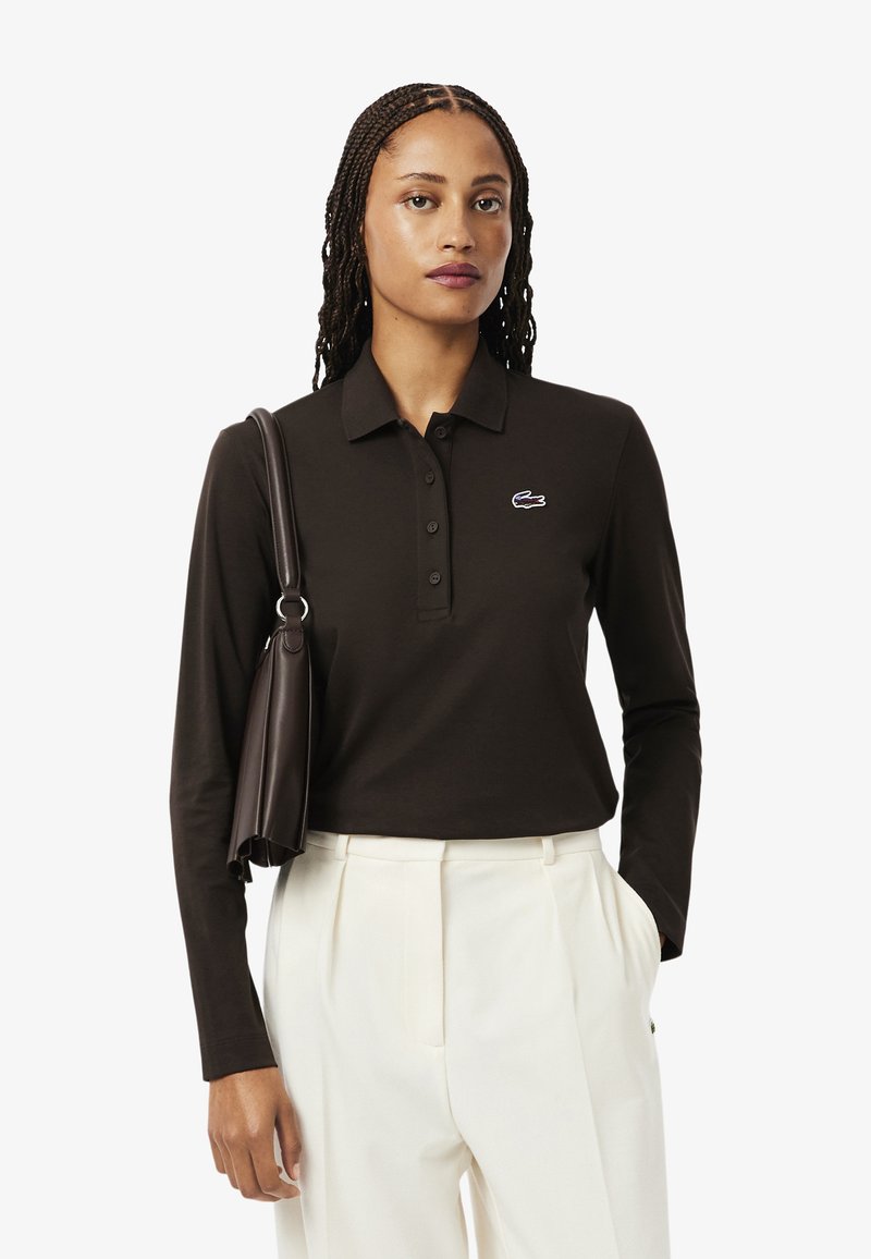 Camisa polo de manga larga marrón oscuro con un tapeta de tres botones y un pequeño logotipo bordado. Combinada con pantalones blancos a medida y un bolso oscuro.