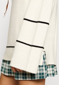 Jersey de canalé crema con rayas negras, combinado con una falda a cuadros verde y blanca. Textura y detalles en el dobladillo notables.