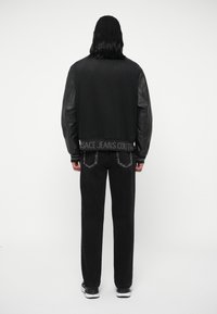 Persona con cabello hasta los hombros que lleva una chaqueta de mangas de cuero negro y jeans negros, de pie de espaldas sobre un fondo simple.