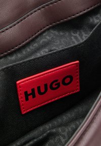 Burgundy nahka sisustus laukussa, jossa on punainen tarra, jossa lukee "HUGO" mustalla tekstillä. Näkyvissä on musta tekstuurihoitokangas.