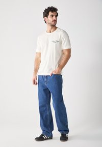 White cotton t-shirt with short sleeves and printed text on the chest, paired with loose blue denim jeans and black sneakers with white stripes.